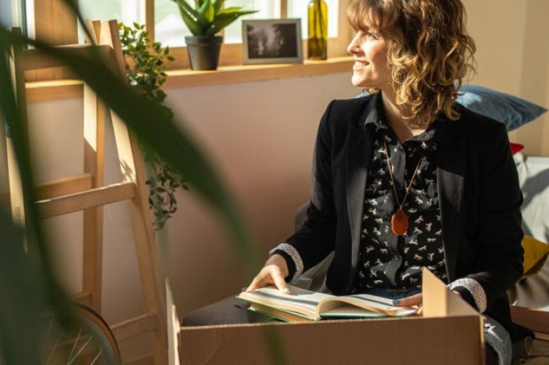 Cheerful attractive woman unpacking cardboard boxes in her new apartment, looking through the window