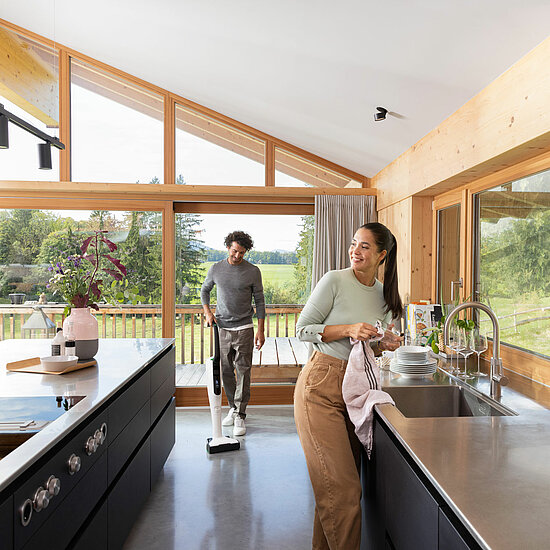 Vorwerk: Photo: A woman dries dishes at a sink while a man vacuums the floor in a modern, sun-drenched kitchen with large windows and a view of a green landscape.